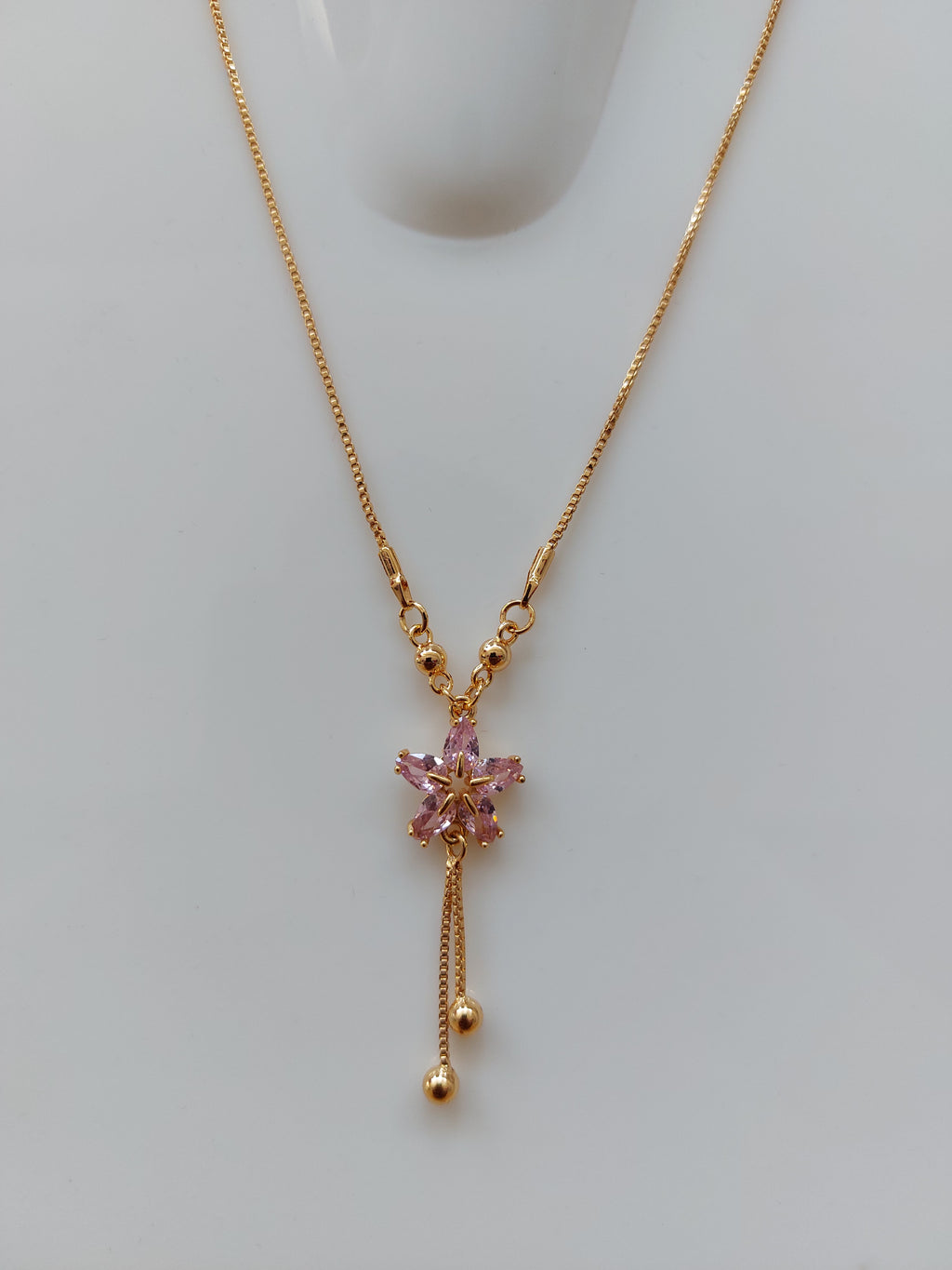 A delicate gold chain necklace with a small pink floral pendant and dangling chain ends, displayed on a light gray surface.