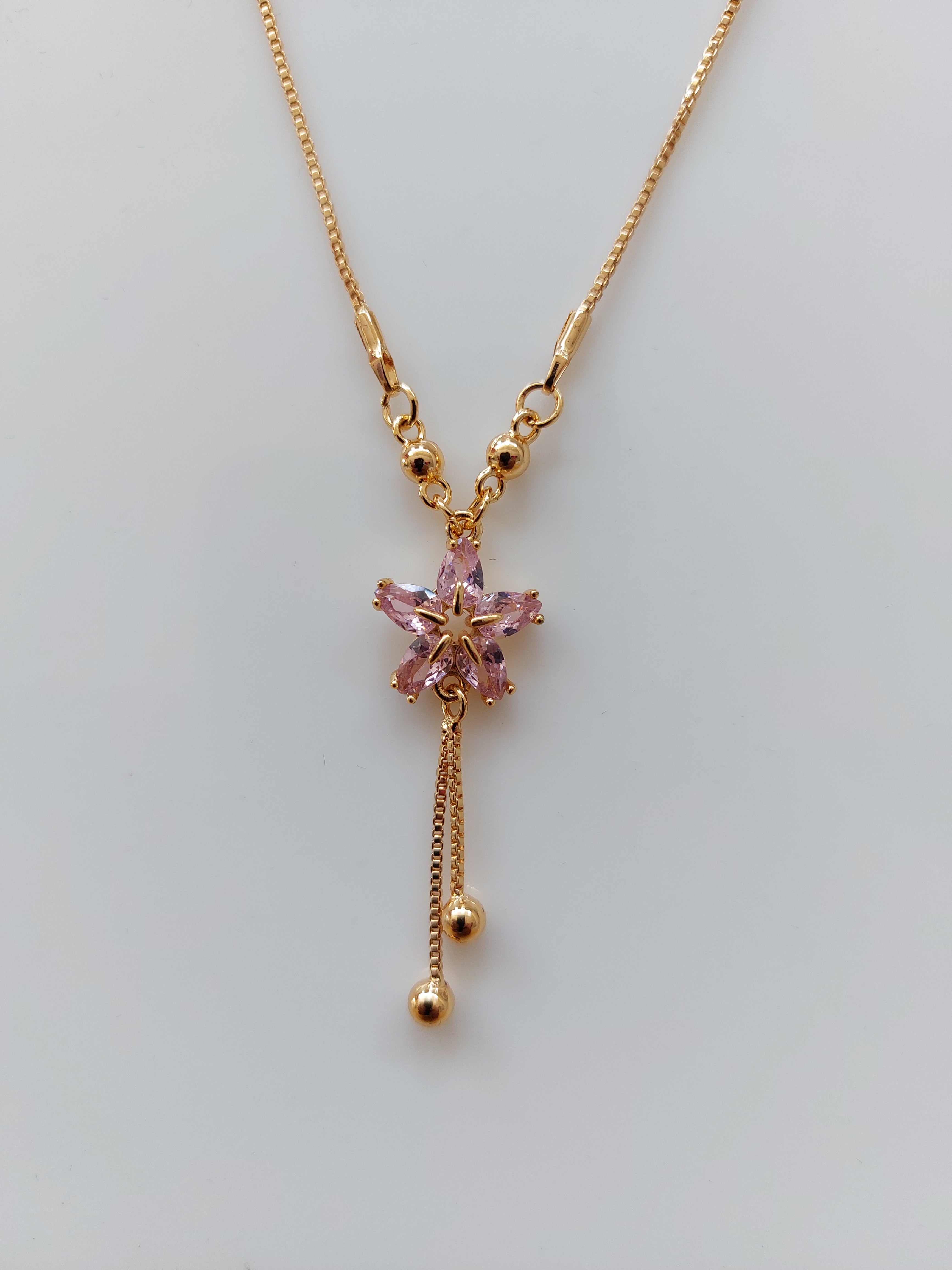 A delicate gold chain necklace featuring a central pink flower-shaped crystal pendant with two dangling chain tails ending in small gold beads, displayed on a light gray background.