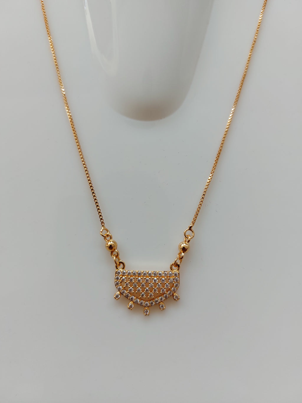 A gold chain necklace with a semi-circular pendant paved with small clear stones and featuring small gold spikes along the curved top edge, displayed on a white mannequin bust against a light gray background.