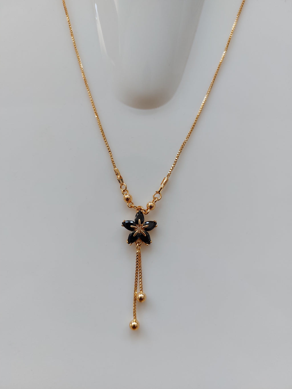 A delicate gold chain necklace with a central black flower charm and small gold bead accents, displayed on a light gray surface.