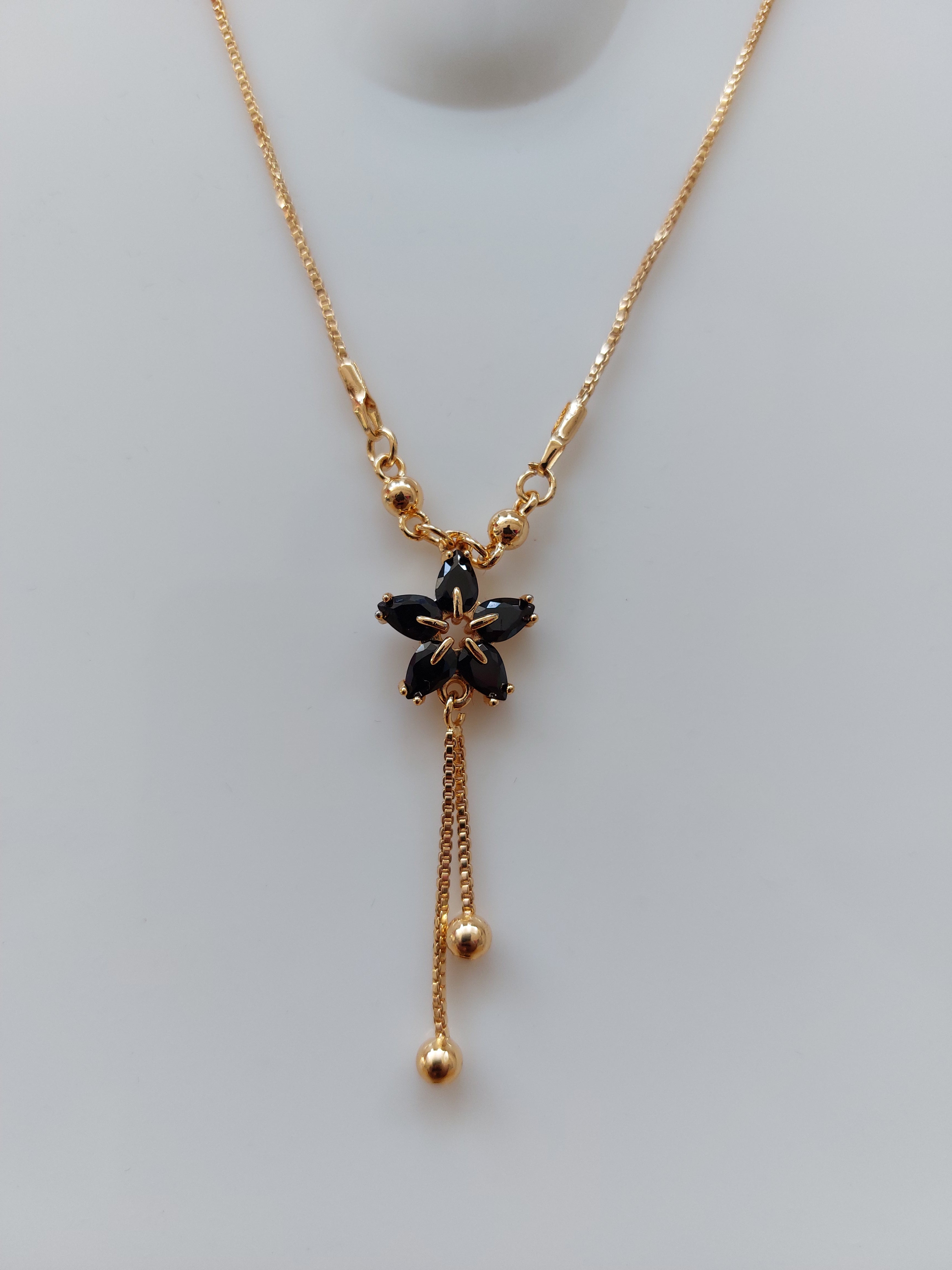 A gold chain necklace featuring a central black flower charm with five petals and two dangling gold chains ending in small beads, displayed on a light gray background.