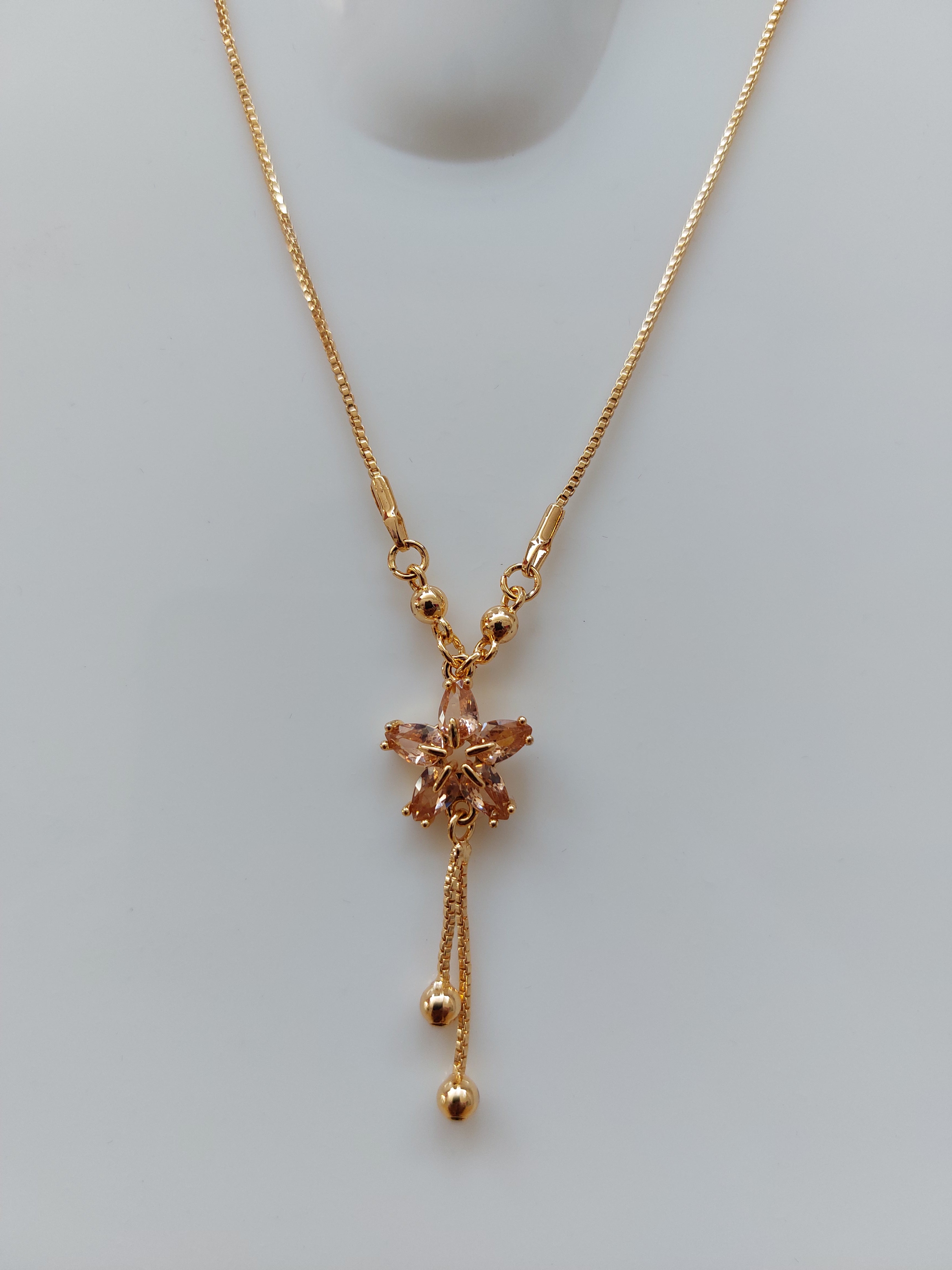 A delicate gold chain necklace with a central floral pendant and two dangling bead chains, displayed on a light gray surface.