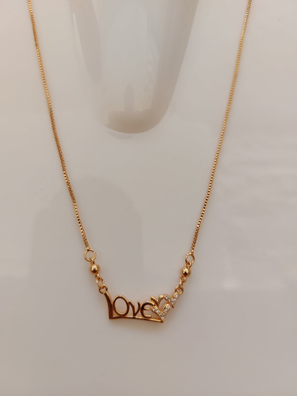 A close-up of a gold chain necklace featuring a decorative clasp with a small clear crystal and a gold heart-shaped accent, displayed on a light off-white background.