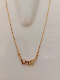 A close-up of a gold chain necklace featuring a decorative clasp with a small clear crystal and a gold heart-shaped accent, displayed on a light off-white background.