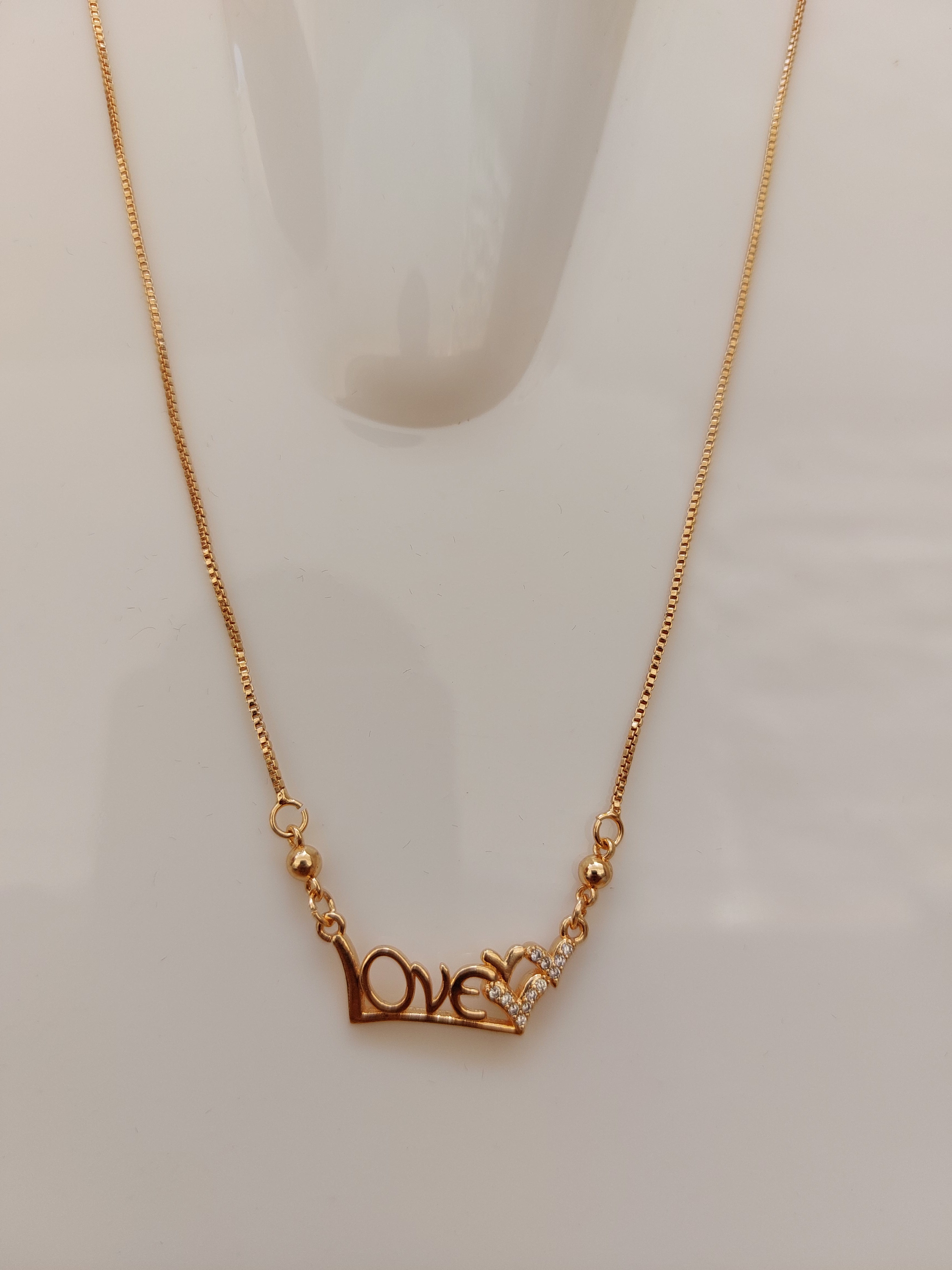 A close-up of a gold chain necklace featuring a decorative clasp with a small clear crystal and a gold heart-shaped accent, displayed on a light off-white background.