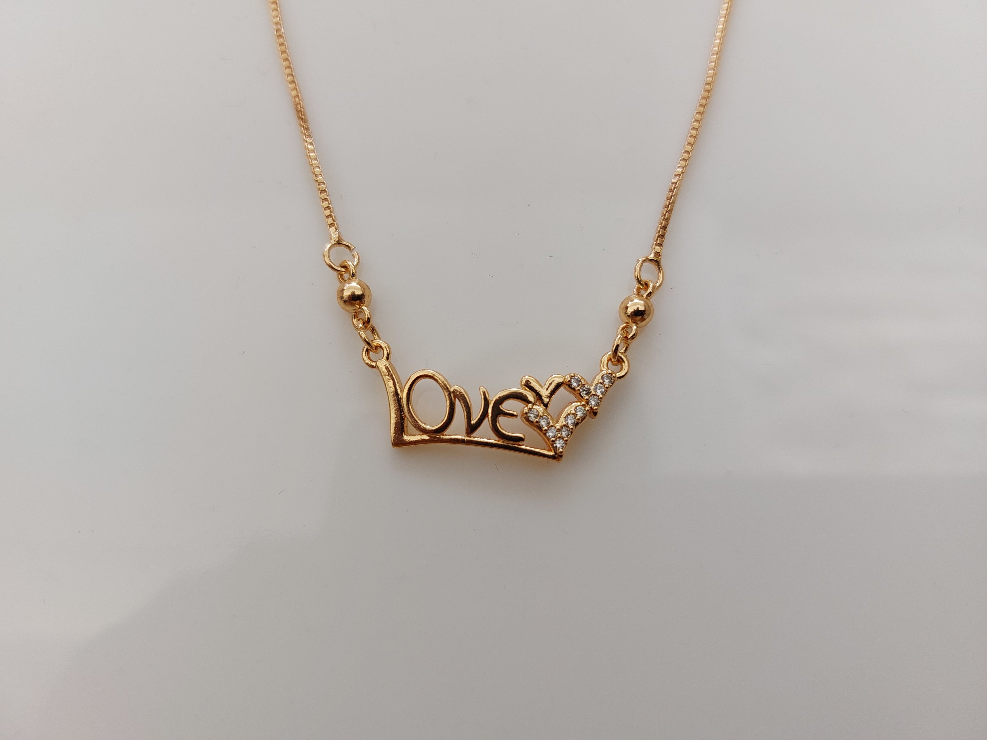 A gold chain necklace featuring a 'LOVE' pendant with a small heart made of clear stones, displayed on a plain light gray background.