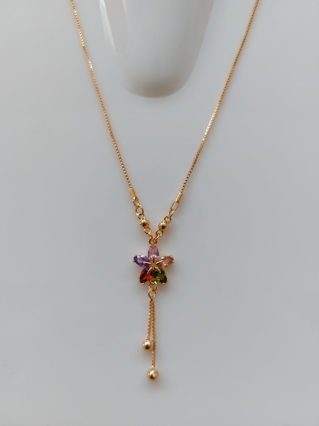 A delicate gold chain necklace with a central floral cluster of small multicolored gemstones, displayed on a light gray surface.