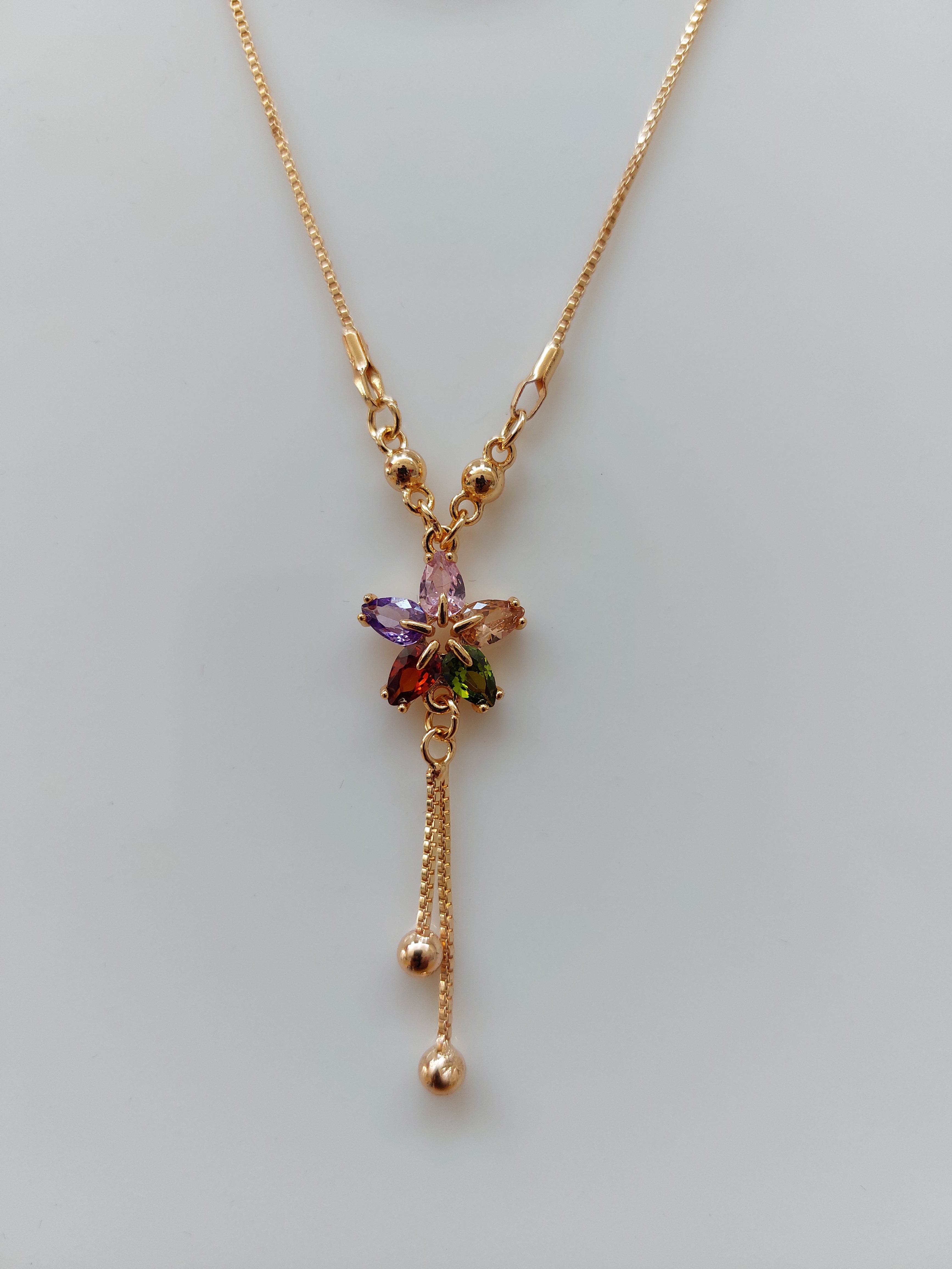 A delicate gold chain necklace featuring a central flower charm with purple, green, and orange enamel details, and two dangling chain ends with small gold beads, shown on a light gray background.
