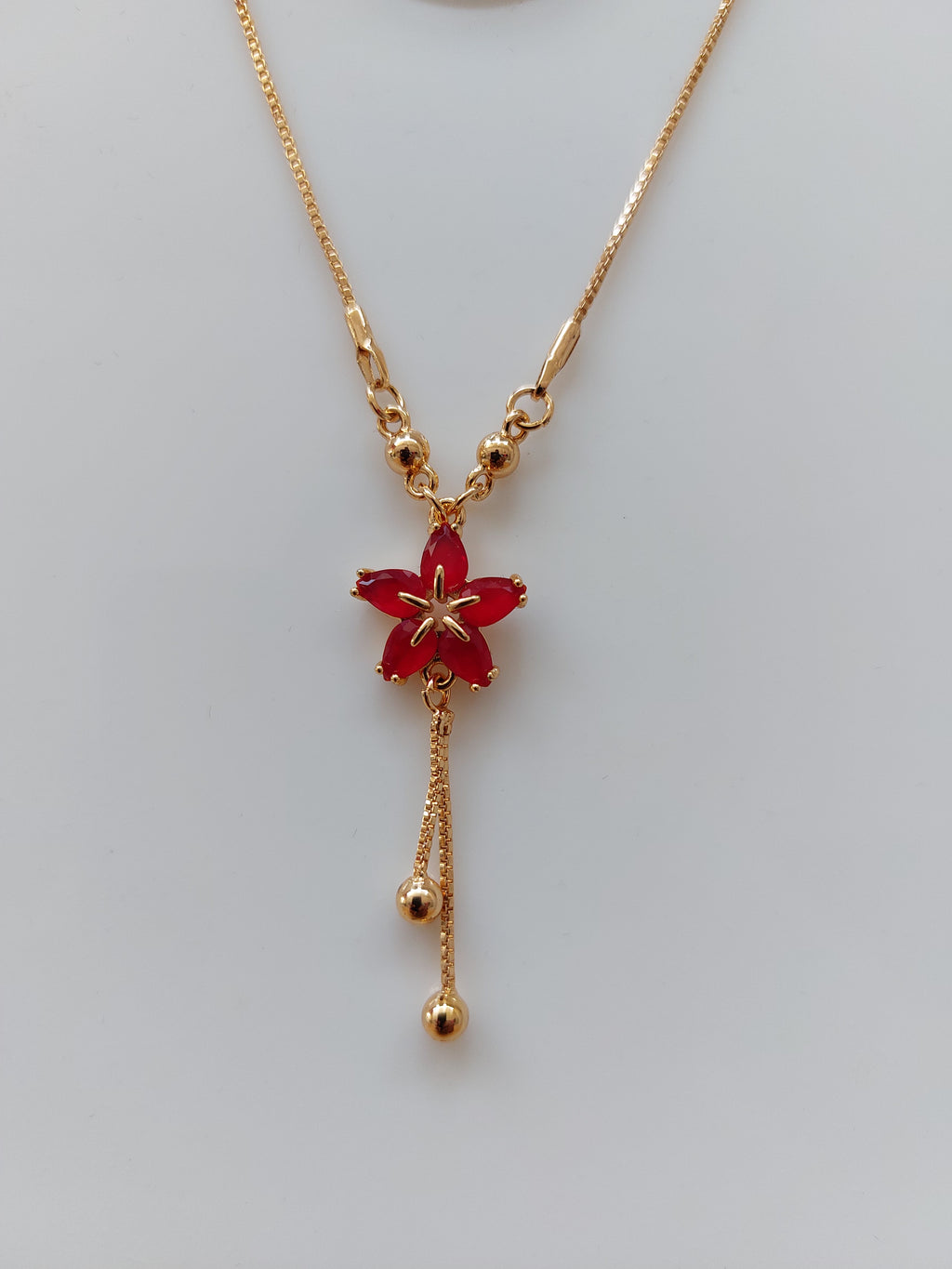 A delicate gold chain necklace featuring a central red flower pendant with gold star details and two dangling gold chain ends with small beads, displayed on a light gray background.