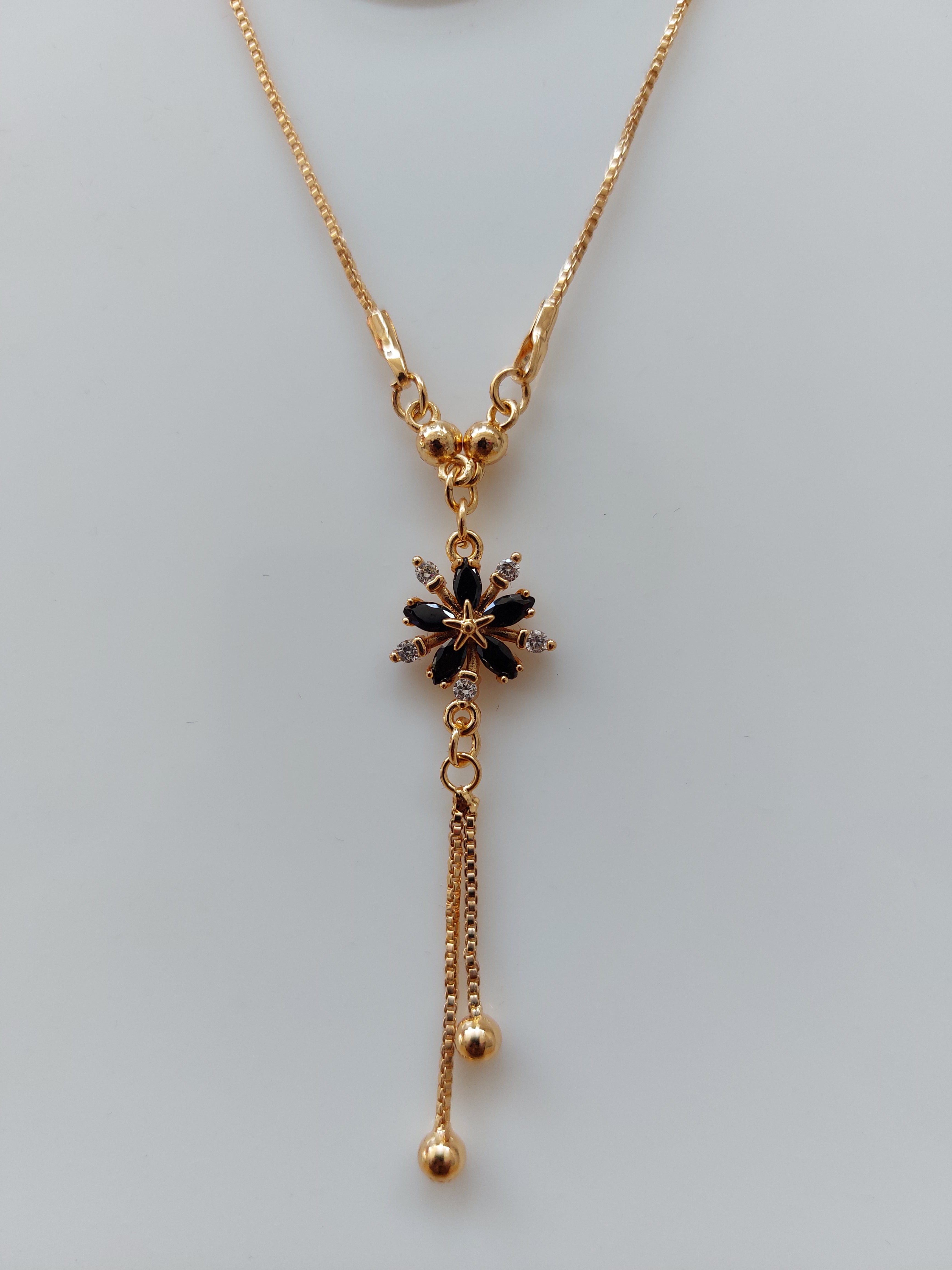 A gold-tone pendant necklace featuring a flower-shaped charm with dark inlay details and two dangling chain tassels, displayed against a plain light gray background.