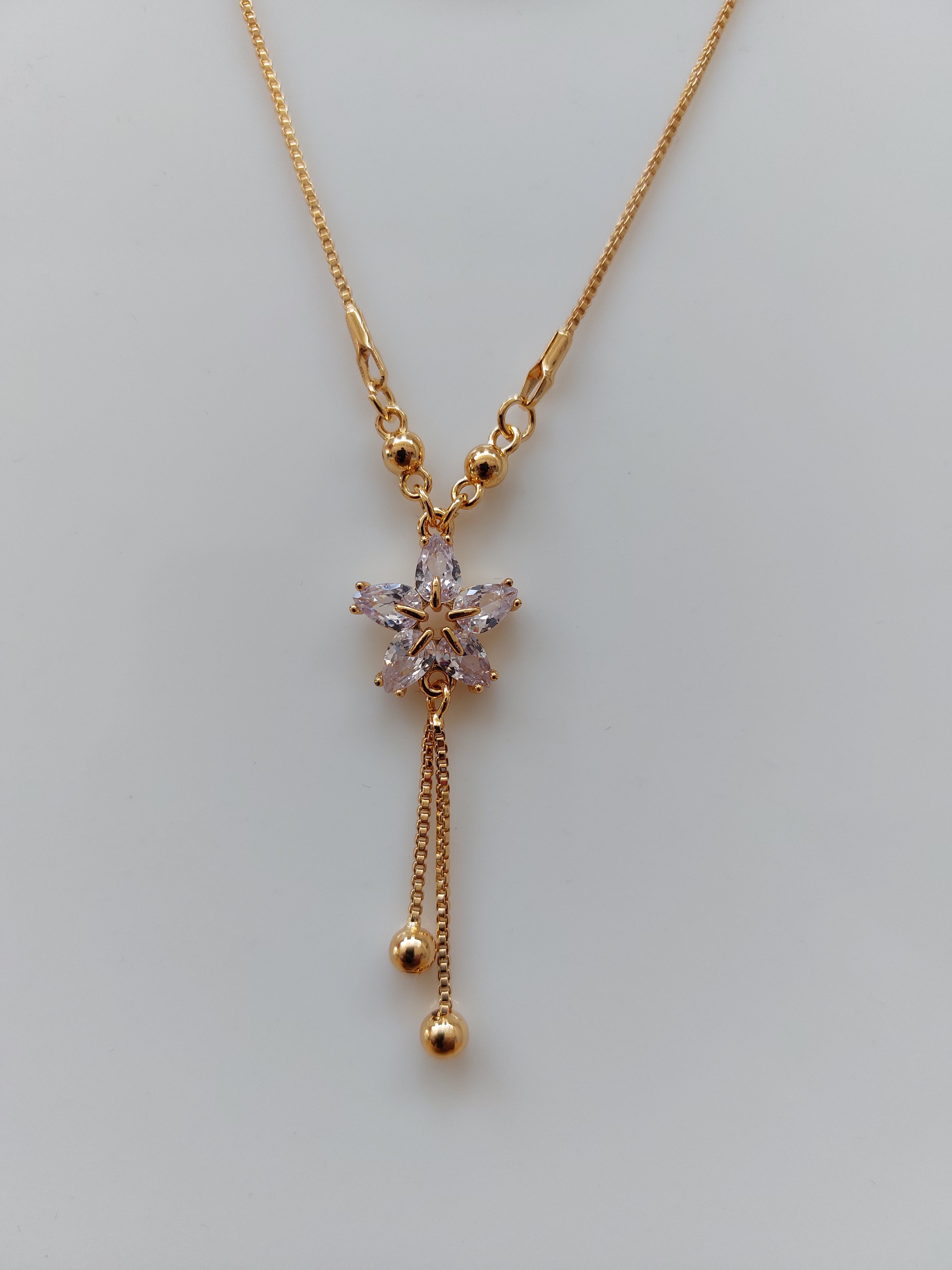 A gold-toned necklace featuring a central flower-shaped crystal pendant with three dangling chain ends, photographed on a plain light gray background.