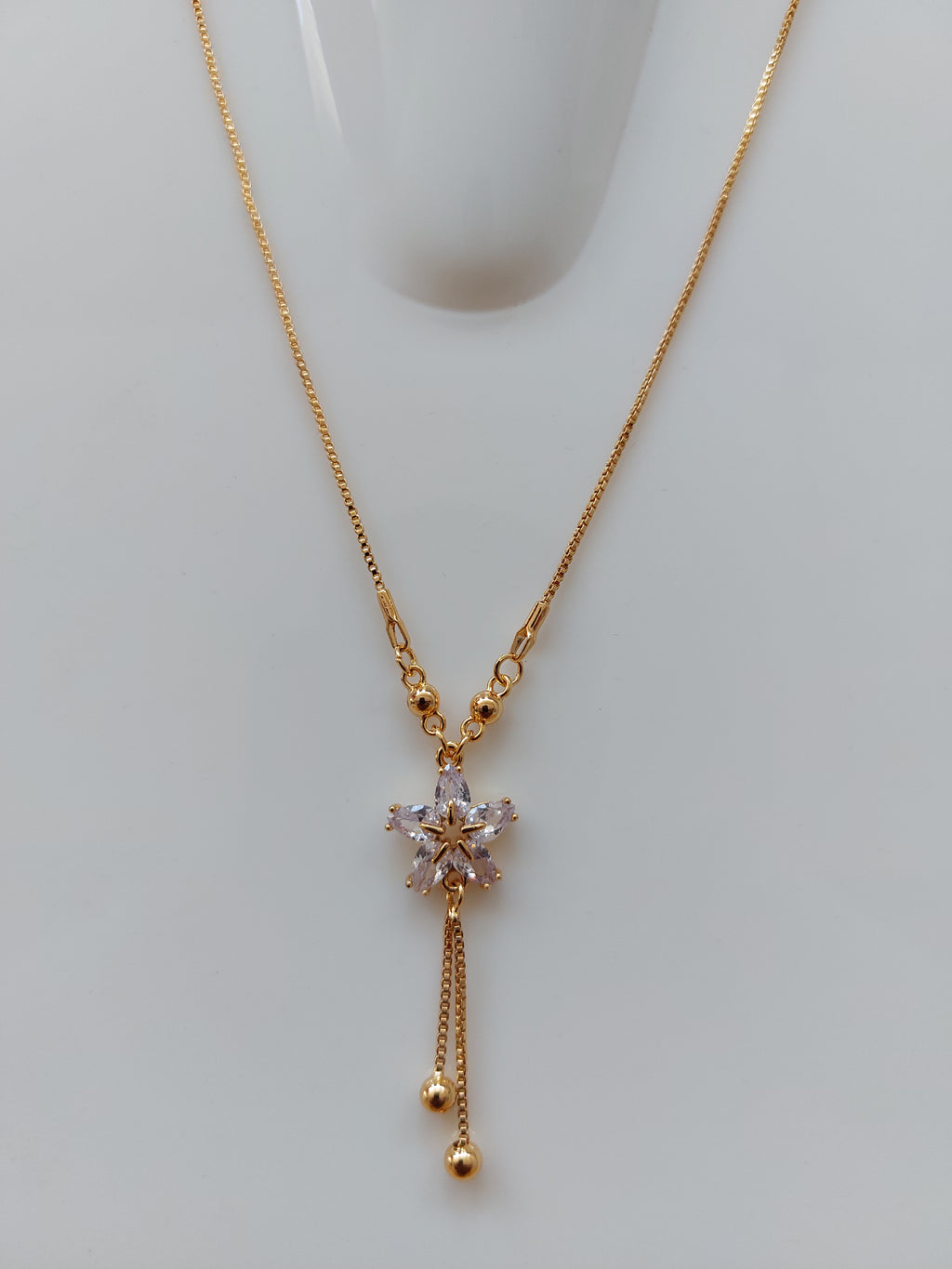 A delicate gold chain necklace with a central floral starburst pendant featuring clear faceted stones, and two dangling chain ends with small gold beads, displayed on a light gray surface.