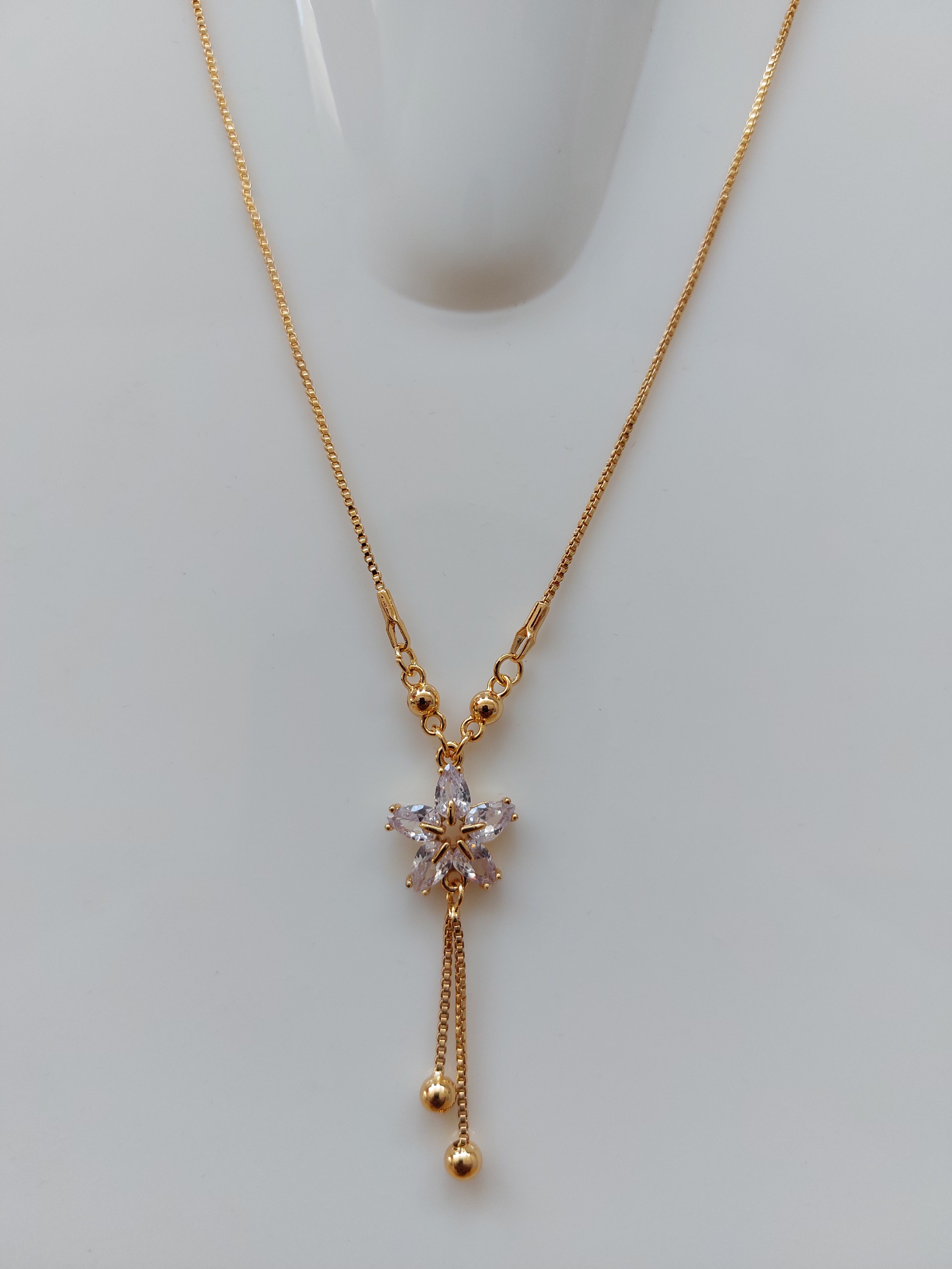 A delicate gold chain necklace with a central floral starburst pendant featuring clear faceted stones, and two dangling chain ends with small gold beads, displayed on a light gray surface.